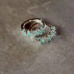 May include: A pair of silver hoop earrings with small, light blue gemstones set around the hoops. The earrings are displayed on a gray, textured surface, with one earring slightly overlapping the other.