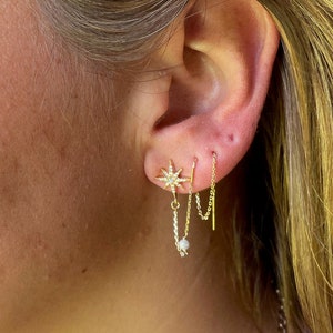 May include: A woman with blonde hair wearing a gold necklace and multiple gold earrings. The earrings include a star-shaped earring with small diamonds, a small pearl earring, and a long chain earring with a small pearl at the end.