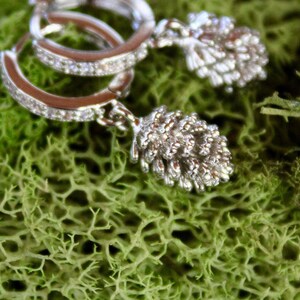 Puede incluir: Pendientes de aro plateados con un diseño de piedras preciosas claras y brillantes. Cada pendiente presenta un pequeño y detallado dije de piña. Los pendientes descansan sobre un lecho de musgo verde vibrante.