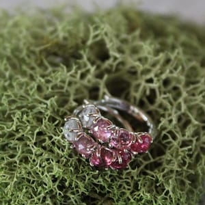 May include: Two silver rings with pink and clear gemstones rest on a bed of green moss. The rings feature a row of small, round gemstones in varying shades of pink and clear. The moss is a vibrant green color.