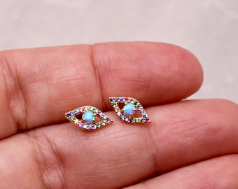 May include: A pair of gold-toned earrings with a blue opal stone in the center of each earring. The earrings are shaped like an eye and are surrounded by colorful gemstones.