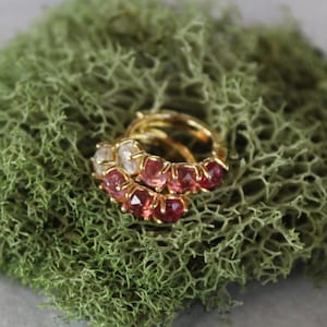 May include: Two gold rings with pink and clear gemstones rest on a bed of green moss. The rings are stacked together, showcasing the gemstones' sparkle. The moss provides a natural, textured backdrop.