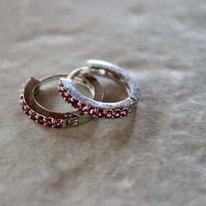 May include: A pair of silver hoop earrings with a row of pink gemstones around the edge.