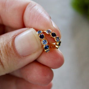 May include: A pair of gold hoop earrings, each featuring a row of blue gemstones. The gemstones are in varying shades, creating a gradient effect. The earrings are held in a hand, highlighting their design and colour.