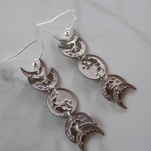 Può includere: Orecchini color argento con un design celeste. Ogni orecchino presenta una serie di fasi lunari, con forme a mezzaluna e luna piena. Gli orecchini sono sospesi a un gancio e sono esposti su una superficie di marmo.
