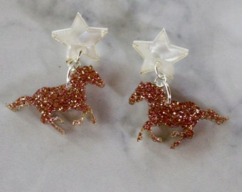 May include: A pair of gold glitter horse earrings with a white star charm hanging from each earring.