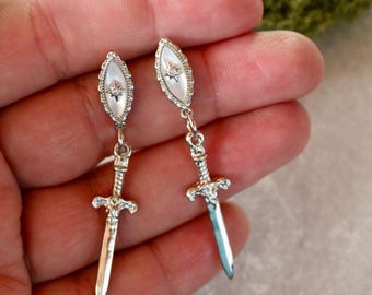 Puede incluir: Un par de pendientes plateados con diseño de espada. Cada pendiente tiene una parte superior ovalada nacarada con un diseño de estrella y un colgante de espada colgante. Las espadas tienen empuñaduras y hojas detalladas. Los pendientes se sostienen en una mano.