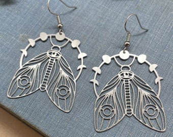 May include: A pair of silver earrings featuring a detailed cutout design of a moth with an eye in the centre. The moth is surrounded by a circle of small dots.