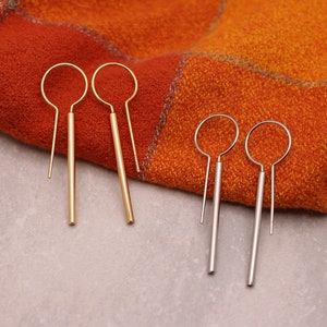 May include: Two pairs of earrings, one gold and one silver, with simple, straight designs. The earrings are on a textured orange and white fabric.
