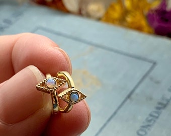 May include: A pair of gold-colored ear cuffs, each featuring an eye design with a central opal-like stone. The eye-shaped design has a detailed, textured border. The ear cuffs are shown against a blurred background.