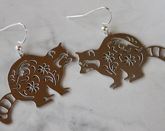 May include: A pair of silver earrings featuring a detailed cutout design of raccoons with floral patterns. The earrings have a simple hook closure.
