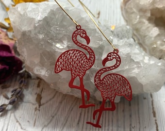 May include: Red flamingo earrings with a filigree design. The earrings have long, gold-colored wires. The earrings are displayed on a white and gray surface.
