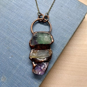 May include: A copper necklace with a pendant featuring three stacked crystals: green, yellow, and purple. The crystals are set in a copper frame and hang from a simple chain.