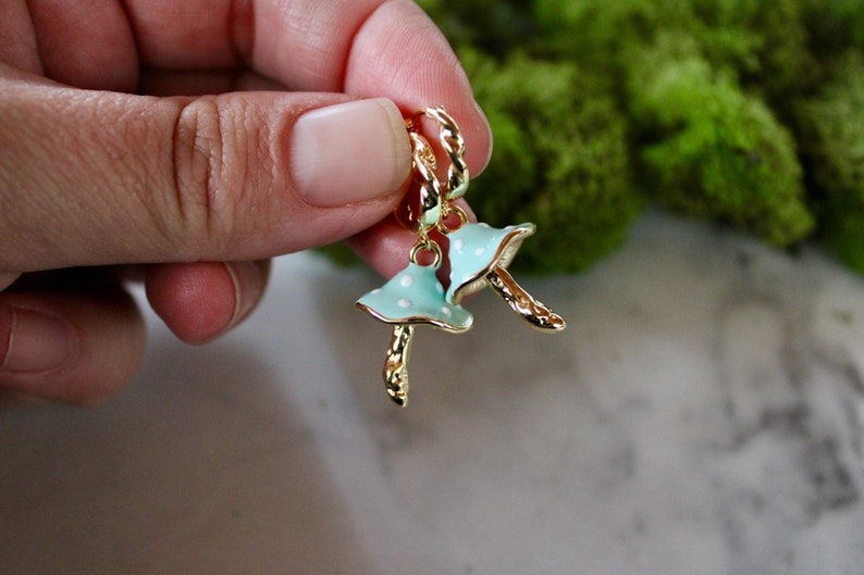 May include: A pair of gold hoop earrings with a blue and white mushroom charm. The mushroom charm has white polka dots.
