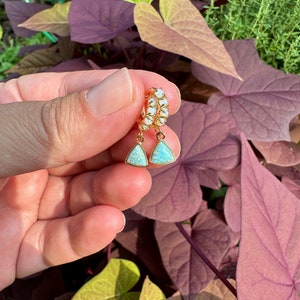 May include: A pair of gold hoop earrings with a teardrop-shaped opal stone dangling from each hoop. The opals are a light blue color with a shimmery finish.