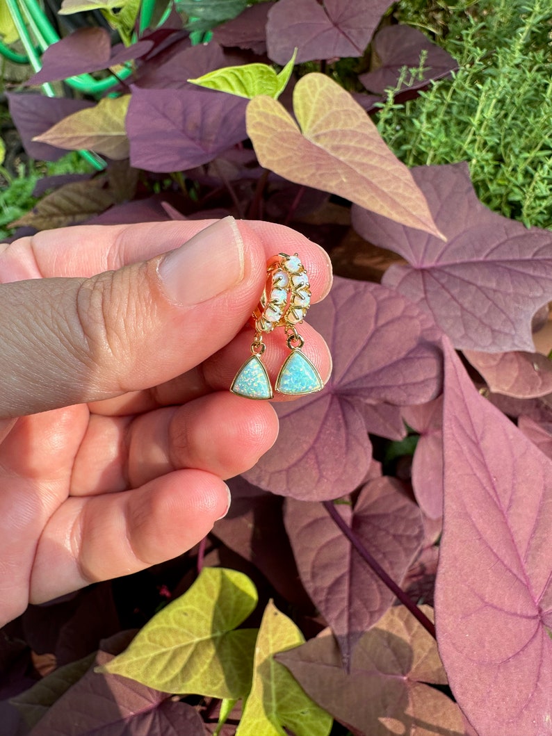 May include: A pair of gold hoop earrings with a teardrop-shaped opal stone dangling from each hoop. The opals are a light blue color with a shimmery finish.