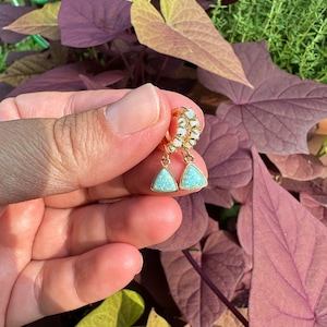 May include: Gold hoop earrings with a teardrop-shaped opal stone dangling from each hoop. The opal stones are a light blue color with a shimmery finish.
