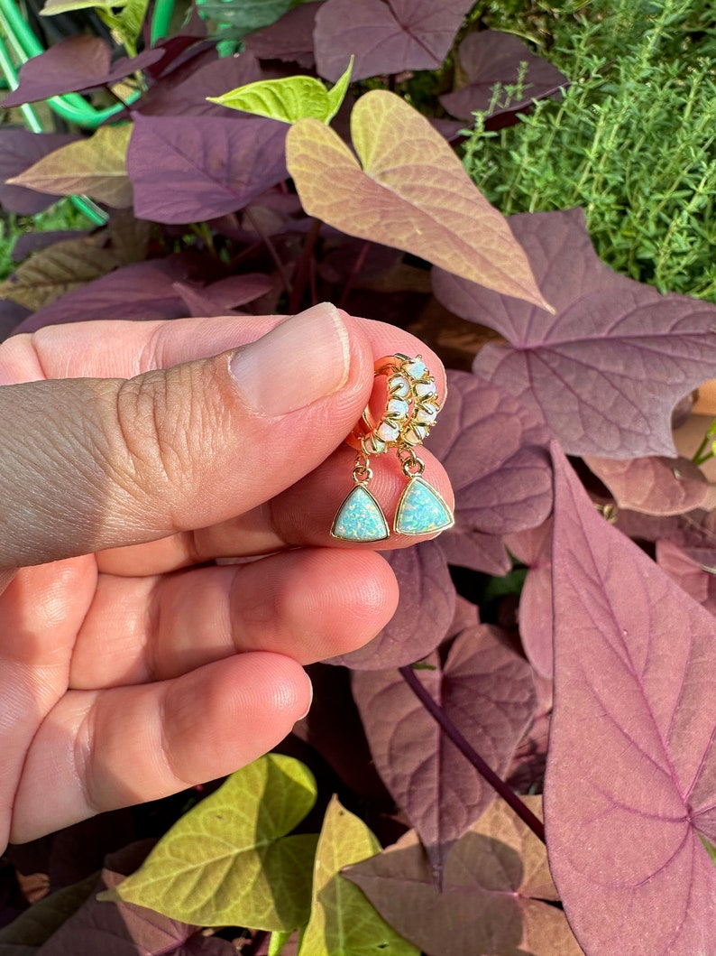 May include: Gold hoop earrings with a teardrop-shaped opal stone dangling from each hoop. The opal stones are a light blue color with a shimmery finish.