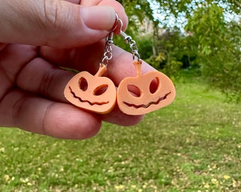 May include: A pair of orange pumpkin-shaped earrings with a carved jack-o'-lantern face. The earrings are made of a lightweight material and have silver hooks.