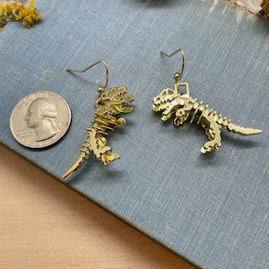 May include: A pair of gold-toned dinosaur skeleton earrings. The earrings are dangling and feature a detailed skeletal design.