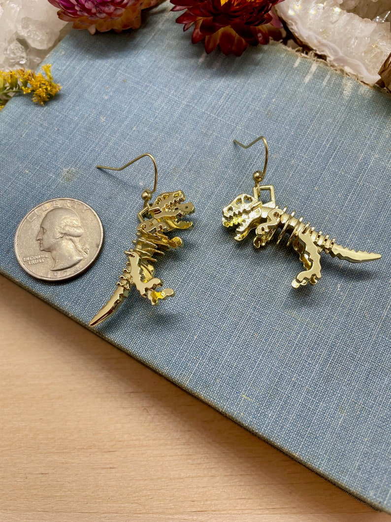 May include: A pair of gold-toned dinosaur skeleton earrings. The earrings are dangling and feature a detailed skeletal design.