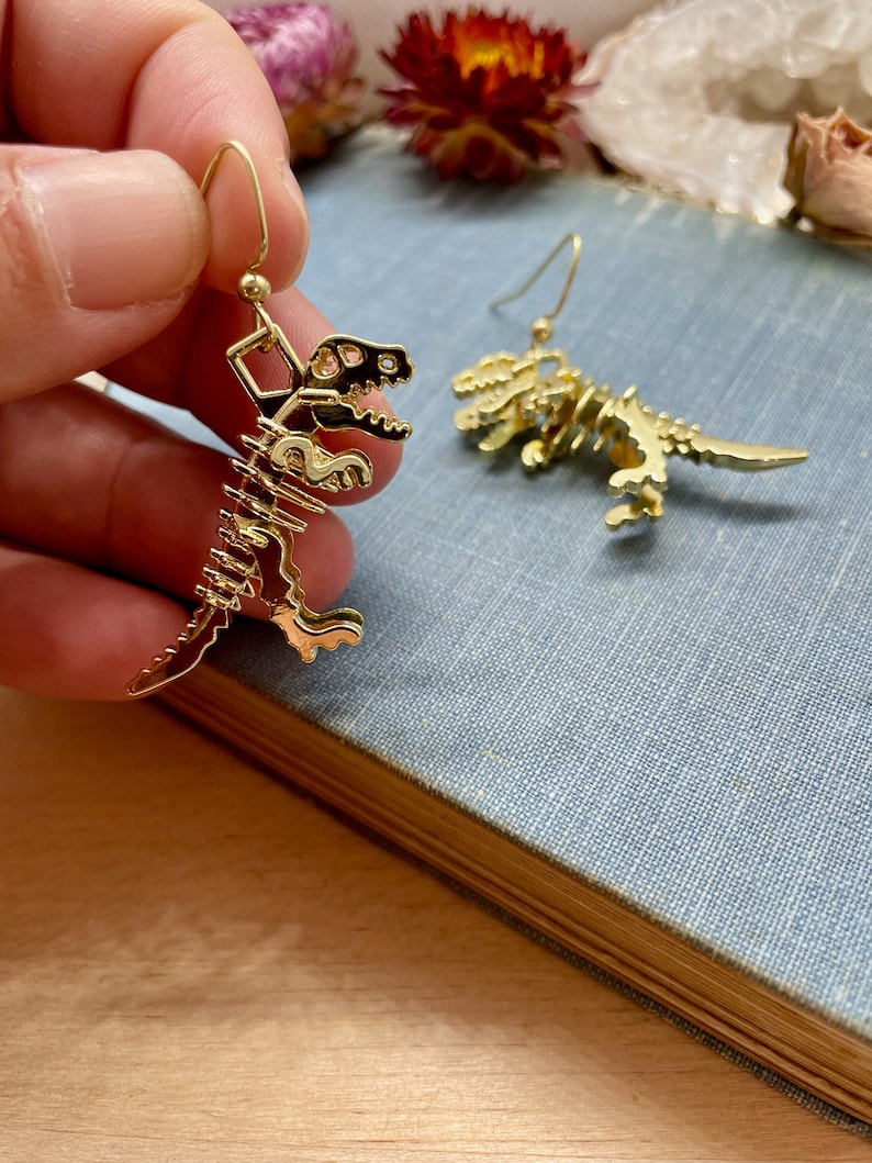 May include: Gold-tone dinosaur skeleton earrings. The earrings are dangling and feature a detailed skeleton design.