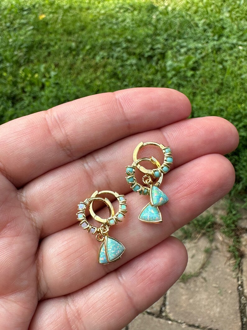 May include: A pair of gold hoop earrings with a dangling triangle-shaped opal gemstone. The hoops are adorned with small white opal stones.