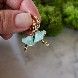 May include: A pair of gold hoop earrings with a blue and white enamel mushroom charm. The mushroom charm has white polka dots.