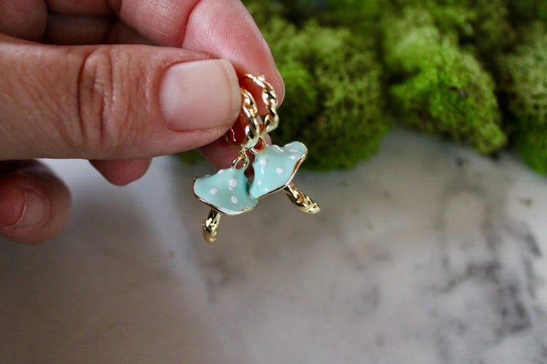 May include: A pair of gold hoop earrings with a blue and white enamel mushroom charm. The mushroom charm has white polka dots.