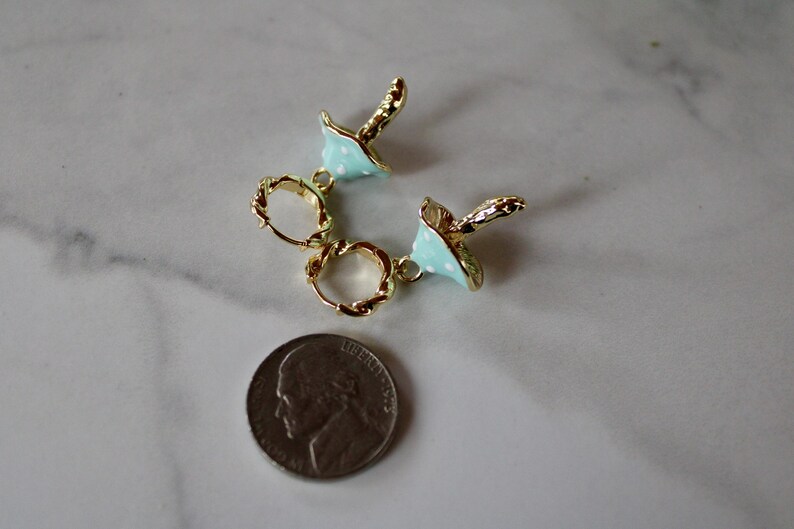 May include: A pair of gold hoop earrings with blue and white enamel mushroom charms. The charms are attached to the hoops with gold jump rings.