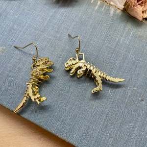 May include: A pair of gold-tone dinosaur skeleton earrings. The earrings are dangling and feature a detailed dinosaur skeleton design.