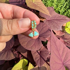 May include: Gold hoop earrings with a turquoise and opal teardrop dangle. The earrings are held in a hand against a backdrop of green and purple leaves.