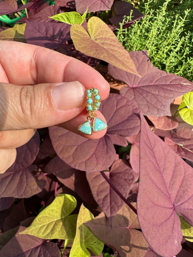 May include: Gold hoop earrings with a turquoise and opal teardrop dangle. The earrings are held in a hand against a backdrop of green and purple leaves.