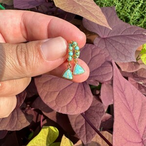 May include: A pair of gold hoop earrings with a turquoise and opal drop. The earrings are held in a hand against a backdrop of green and purple leaves.