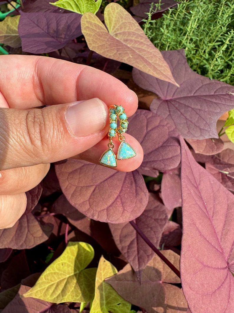 May include: A pair of gold hoop earrings with a turquoise and opal drop. The earrings are held in a hand against a backdrop of green and purple leaves.