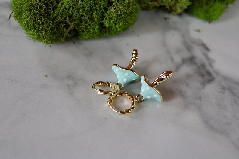 May include: A pair of gold hoop earrings with a light blue mushroom charm with white polka dots hanging from each hoop.