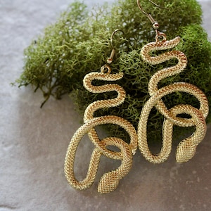May include: Gold-colored snake-shaped earrings with a textured surface. The earrings are displayed on a bed of green moss against a gray background. The earrings have a hook closure.