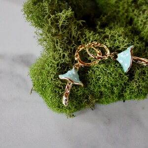 May include: A pair of gold chain earrings with blue and white mushroom charms. The earrings are on a bed of green moss.