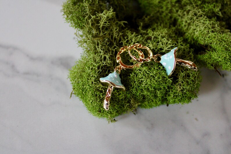 May include: A pair of gold chain earrings with blue and white mushroom charms. The earrings are on a bed of green moss.