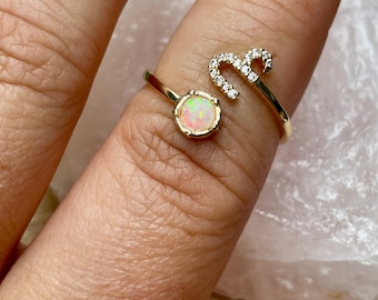 May include: A gold-colored ring with a round, iridescent opal stone and a curved design. The ring features a small cluster of clear, sparkling stones. The ring is worn on a finger.