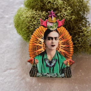 May include: A wooden Frida Kahlo portrait pin. The pin depicts Frida Kahlo with a sunburst halo, a green dress, and a butterfly. The pin is set against a backdrop of green moss.