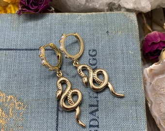 May include: Gold snake hoop earrings with clear cubic zirconia stones. The earrings are dangling from the hoops and have a snake design.