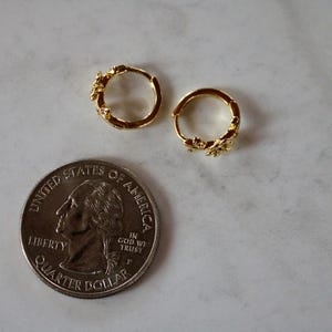 May include: A pair of gold hoop earrings with a floral design. The earrings are small and delicate, and they are shown next to a US quarter for scale.