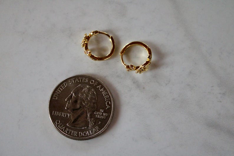 May include: A pair of gold hoop earrings with a floral design. The earrings are small and delicate, and they are shown next to a US quarter for scale.