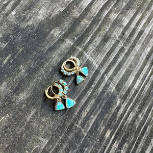 May include: A pair of gold hoop earrings with turquoise triangle charms. The earrings are on a dark wooden surface.