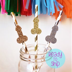 May include: Three decorative straws with glitter penis toppers in gold, silver, and bronze. The straws are striped with gold and white, and black and white. The straws are in a clear glass jar. Colorful streamers are in the background.