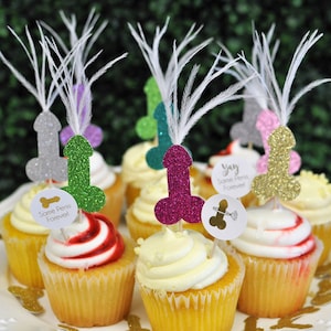 bachelorette party cupcakes are adorned with glitter penis cupcake toppers sprouting feathers from the tops. a bride is pictured in shocked laughter beside the cupcakes.