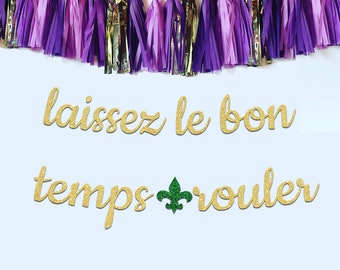 May include: Gold glitter letters spelling "laissez le bon temps rouler" with a green fleur-de-lis symbol. The letters are hanging from a purple and gold tassel garland.