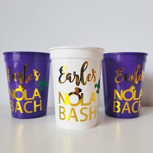 May include: Three purple plastic cups with gold lettering that reads "Earles' Nola Bash" and "Earles' Nola Bach".