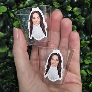 May include: Two clear plastic shot glasses, each featuring a custom sticker of a smiling person with long brown hair wearing a white bridal veil. The small, transparent cups are designed for serving beverages at events or parties.